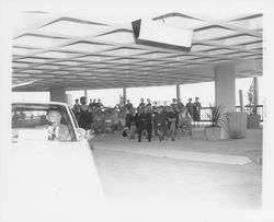 A man drives a white Plymouth Fury while a model displays a black dress behind him in a fashion show at dedication of parking garage at 3rd and D, Santa Rosa, California, 1964 (Digital Object)